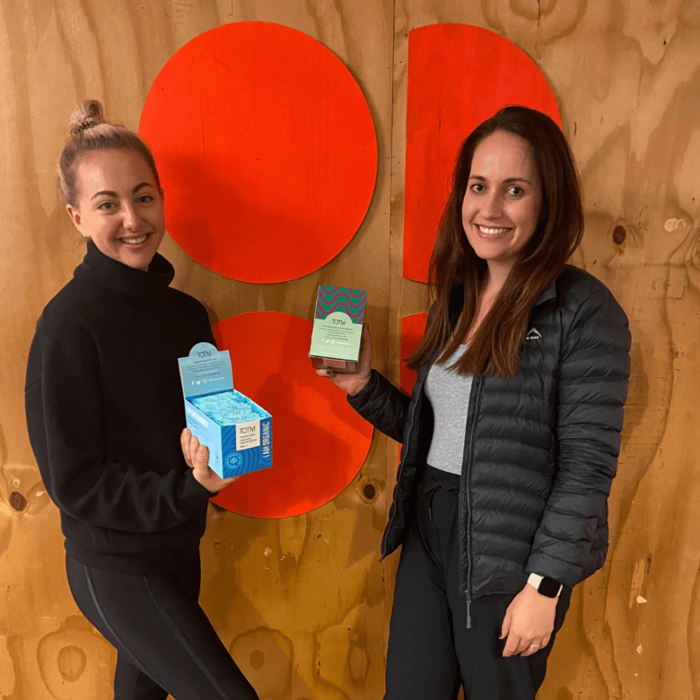 Two members from the Sweaty Betty team standing against a woodchip board with the Sweaty Betty logo. The employees are holding up free period care products as they are signed up to TOTM's period positive workplace scheme.