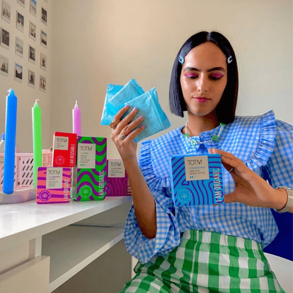 Girl with short black hair in a blue gingham blouse and green check trousers is holding a box of TOTM pads and loose pads in her hand. She's surrounded by TOTM period care as a fan of this award-winning period care brand.