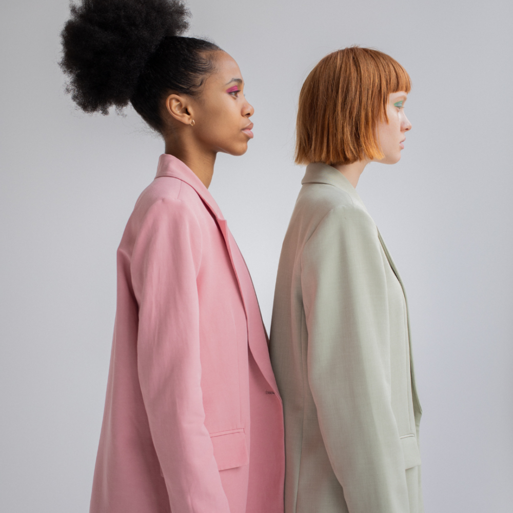 Two women facing right in suits. One is in a pink suit and one is in a pale green/cream suit. They are serious as this represents the negative impact of period stigma in work and lack of free period care products.