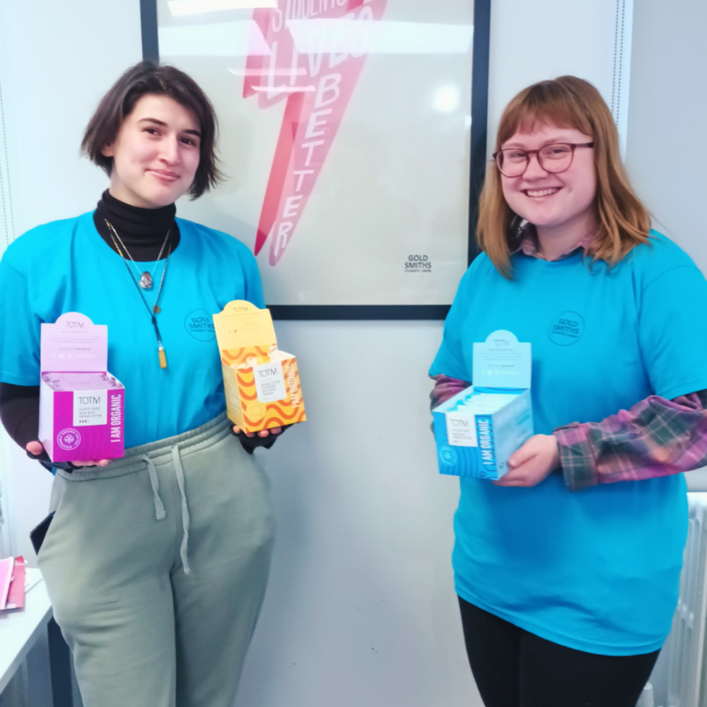 Two students from Goldsmith's University smiling in a bathroom holding TOTM period products as Goldsmiths are providing free period care