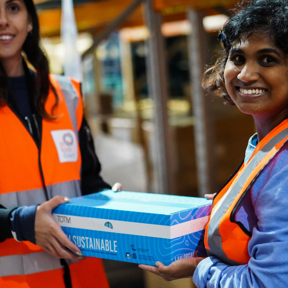 Two women who work for In Kind Direct dressed in fluorescent orange vests holding a box of TOTM pads which will be distributed to their network to support period dignity and fight period poverty in the UK.