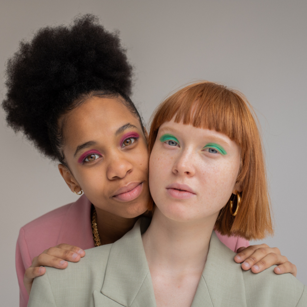 Two women wearing pastel coloured suits facing the camera with colourful eyeshadow